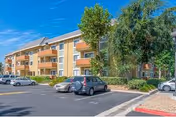 Three-story orange-brick senior living building with balconies, trees, and a parking lot.