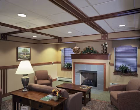 A cozy living room area with a fireplace in the center, flanked by two windows with blinds and potted plants on the sills. The room features comfortable upholstered chairs and a sofa, a wooden coffee table with magazines, a side table with a lamp, and decorative items on the fireplace mantel. The ceiling has recessed lighting and wooden beams.