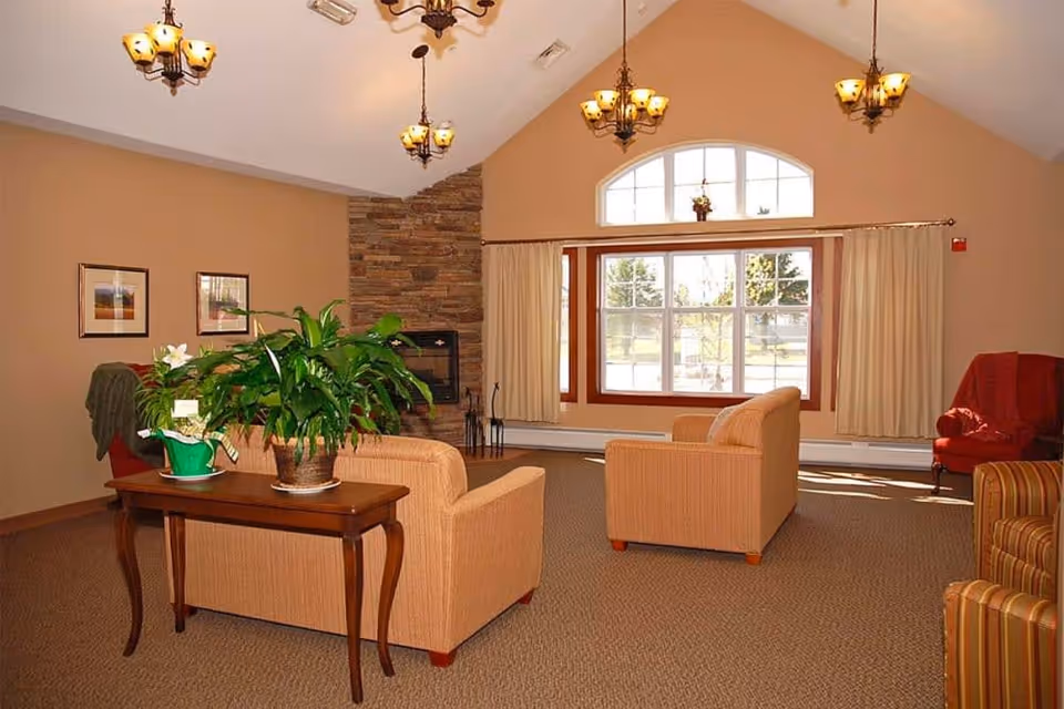 A cozy senior living common area with beige walls and carpet, featuring two beige armchairs facing a large window with cream curtains. A wooden table with green potted plants is placed behind one armchair. The room has a stone fireplace on one wall and several hanging light fixtures from the vaulted ceiling. Additional seating includes a red armchair and a striped armchair.