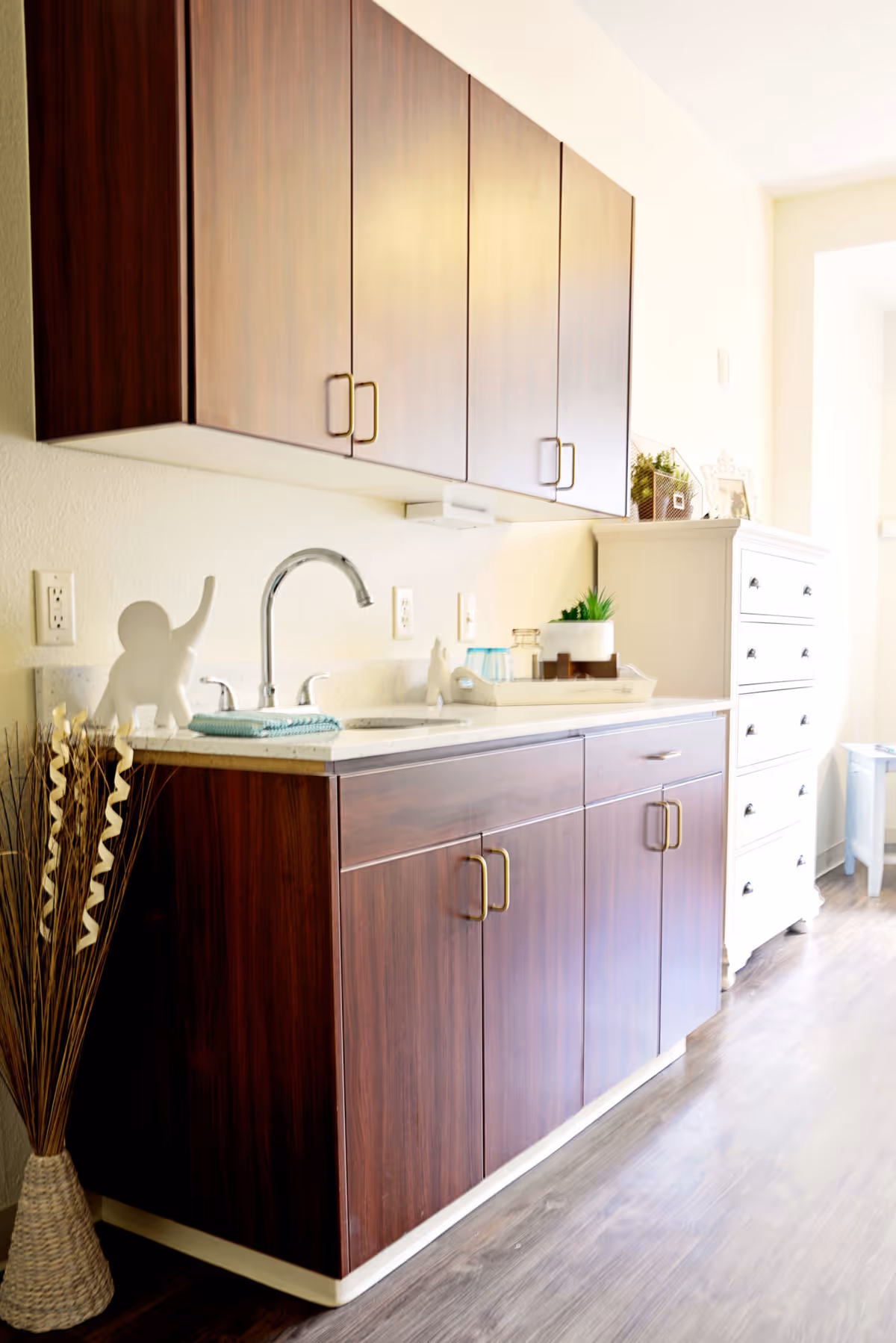 A bright interior space featuring dark wood cabinets with brass handles, a countertop with a sink and faucet, decorative items including a white elephant figurine, a tray with cups and plants, and a white chest of drawers in the background on a wooden floor.