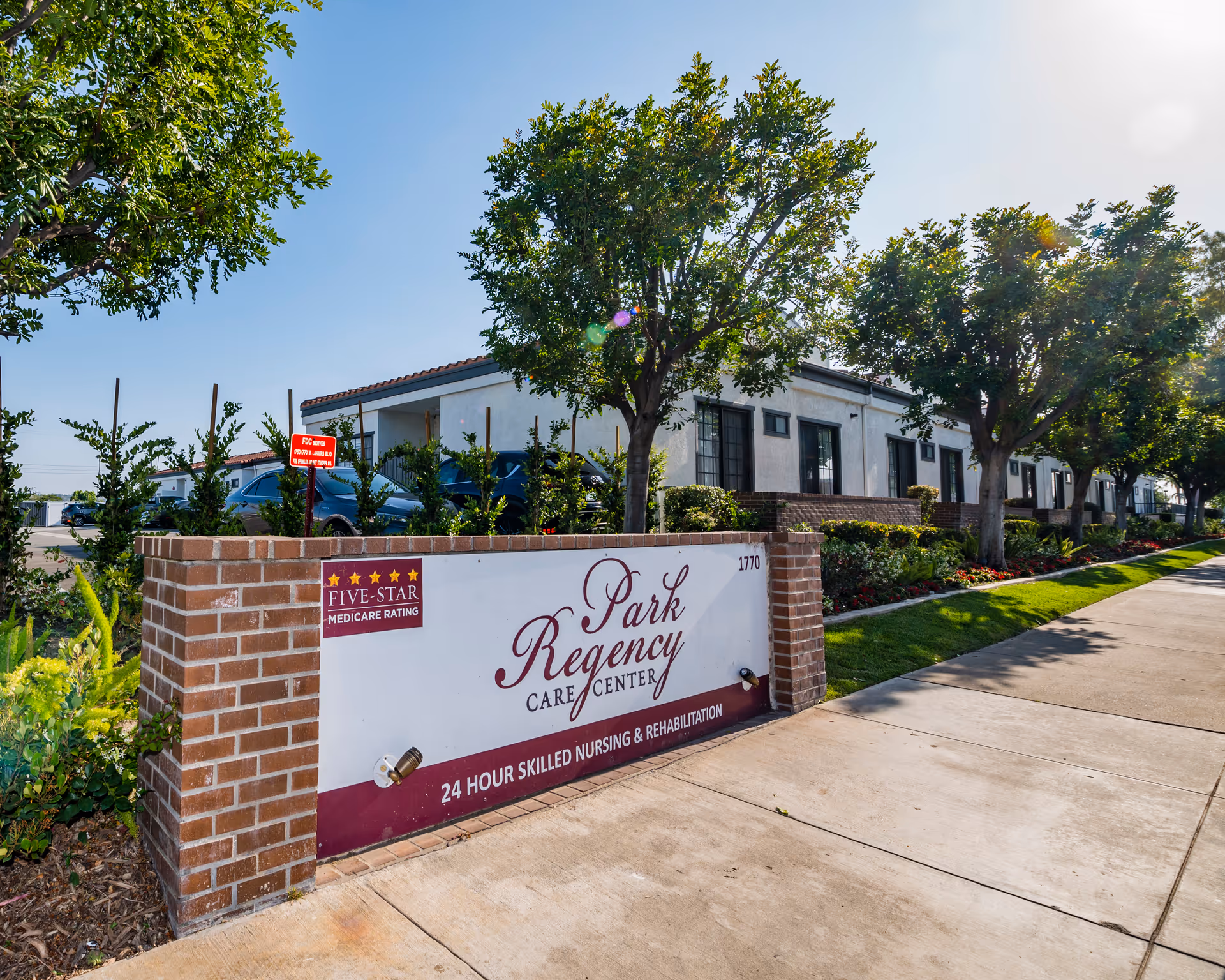Front sign reading 'Park Regency Care Center' on a low brick wall with the facility building, trees, and sidewalk in the background.