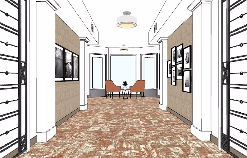 A hallway with patterned carpet flooring, beige walls adorned with framed black and white photographs, and two orange chairs with a small round table between them positioned near large windows at the end of the hall.
