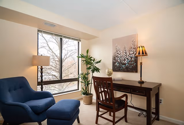 A cozy interior room with a large window showing bare tree branches outside. Inside, there is a blue upholstered armchair with a matching ottoman, a tall floor lamp with a beige shade, a wooden desk with a matching wooden chair, a small globe on the desk, a potted plant, and a wall painting featuring abstract floral designs. The room has beige walls and carpeted floor.
