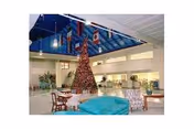 Spacious indoor common area with a high ceiling and large windows. The room features a tall decorated Christmas tree in the center, surrounded by various seating arrangements including chairs and sofas. The ceiling has a blue glass section with hanging flags. The area appears bright and welcoming.