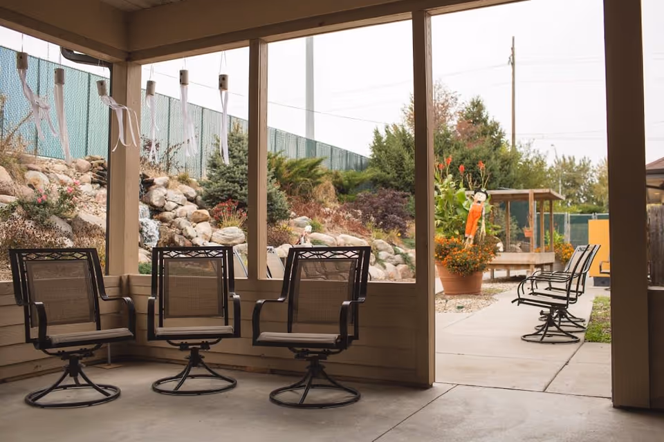 Covered outdoor seating area with several metal mesh chairs arranged around the perimeter. Beyond the covered area, there is a concrete pathway with more chairs and potted plants. The background features a rock garden with various shrubs and trees, and a green fence along the top of the slope.