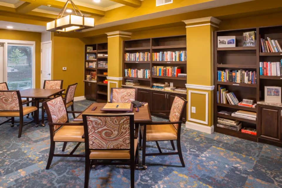 A cozy reading or activity room with several wooden tables and chairs featuring patterned upholstery. The room has mustard yellow walls with white trim and built-in dark wood bookshelves filled with books and magazines. A large window with blinds lets in natural light, and a modern chandelier hangs from the ceiling. The carpet has a blue and yellow abstract pattern.