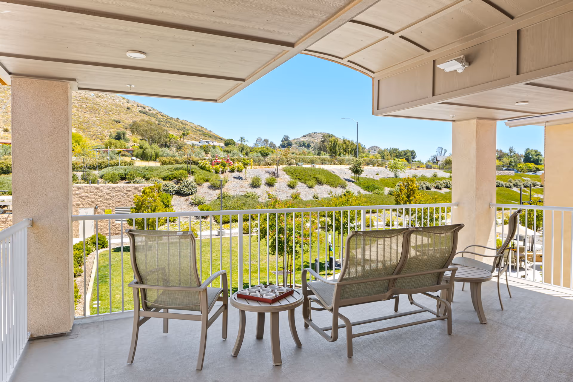 Covered outdoor patio area with metal chairs and a small table, overlooking a landscaped garden with shrubs, trees, and hills in the background under a clear blue sky.