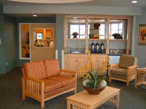 A cozy sitting area in a senior living facility with two wooden armchairs and a matching loveseat upholstered in patterned fabric. A wooden coffee table with a potted plant featuring yellow flowers is in the center. Behind the seating area is a wooden cabinet with a countertop holding coffee dispensers and cups, and shelves with decorative plants. A window and door lead to another room where people are visible.