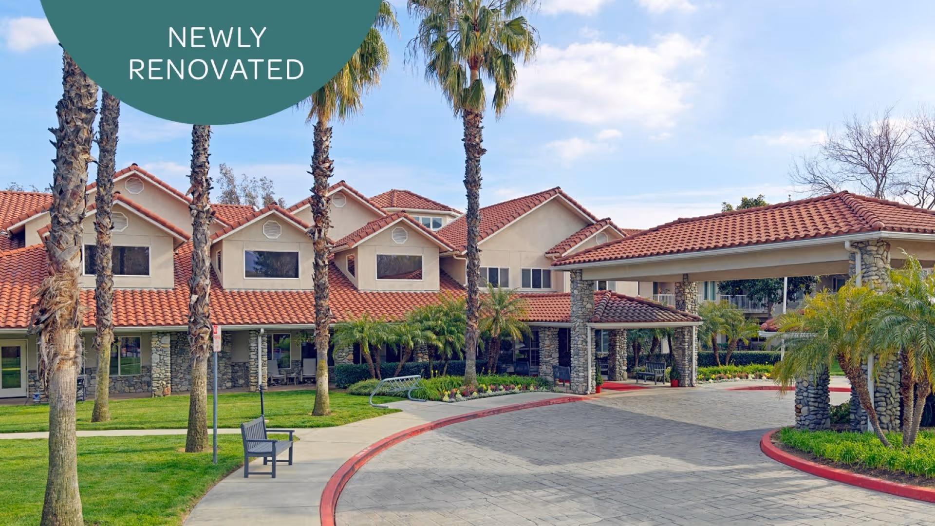 Front entrance of a Mediterranean-style senior living building with red tile roofs, palm trees, and a circular driveway under a covered porte-cochère.