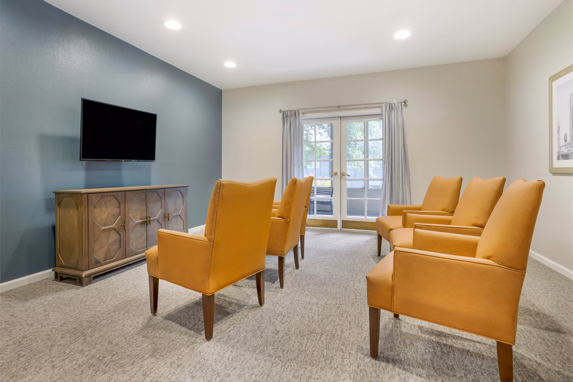 A small common room with mustard-yellow armchairs facing a wall-mounted TV and glass double doors.