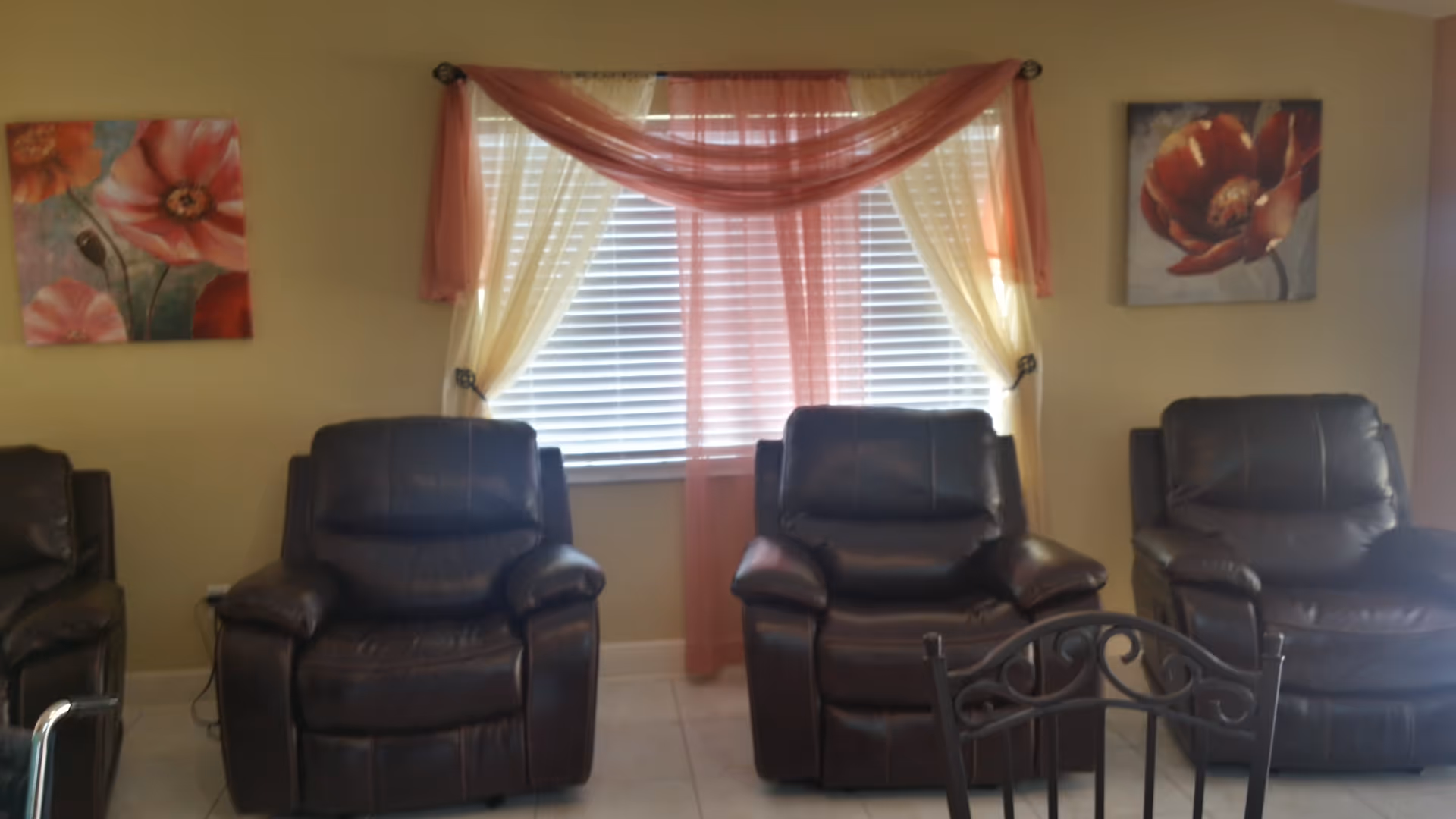 A room with three dark brown leather recliner chairs arranged in front of a window with white blinds and sheer pink and cream curtains. On the wall above the chairs are two floral paintings with red and pink flowers. The floor is tiled and part of a black metal chair is visible in the foreground.