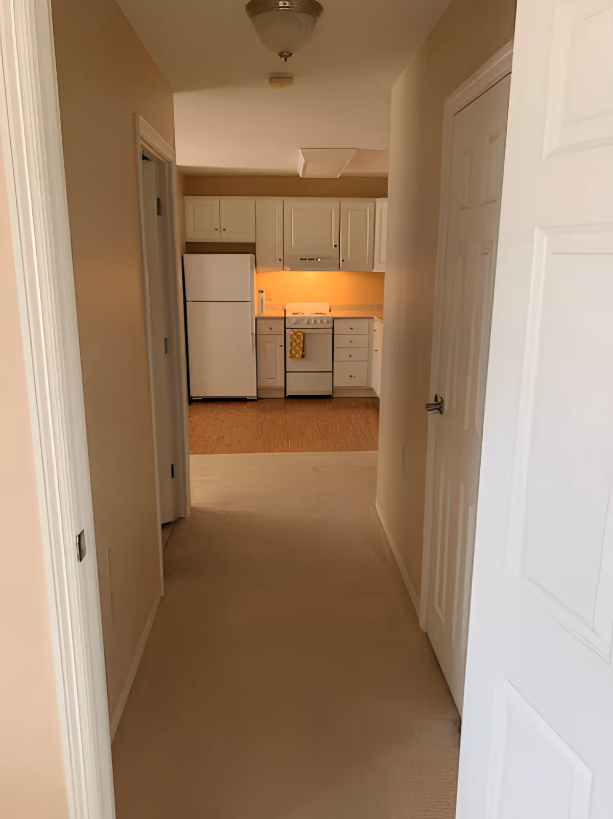 View down a hallway leading to a small kitchen with white cabinets, a white refrigerator, and a white stove with an orange towel hanging on the handle. The hallway has beige walls and carpeted flooring, with a closed white door on the right and an open doorway on the left.