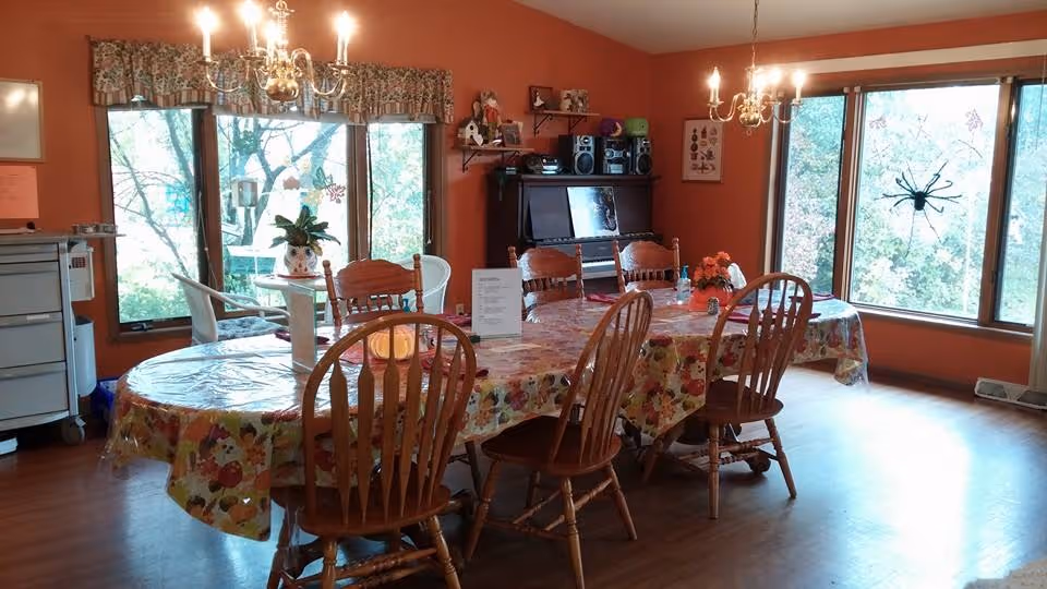 A dining room with orange walls and large windows letting in natural light. There is a long table covered with a floral tablecloth and surrounded by wooden chairs. Two chandeliers hang from the ceiling above the table. A piano and some shelves with decorations are against the far wall. A large decorative spider is attached to one of the windows.