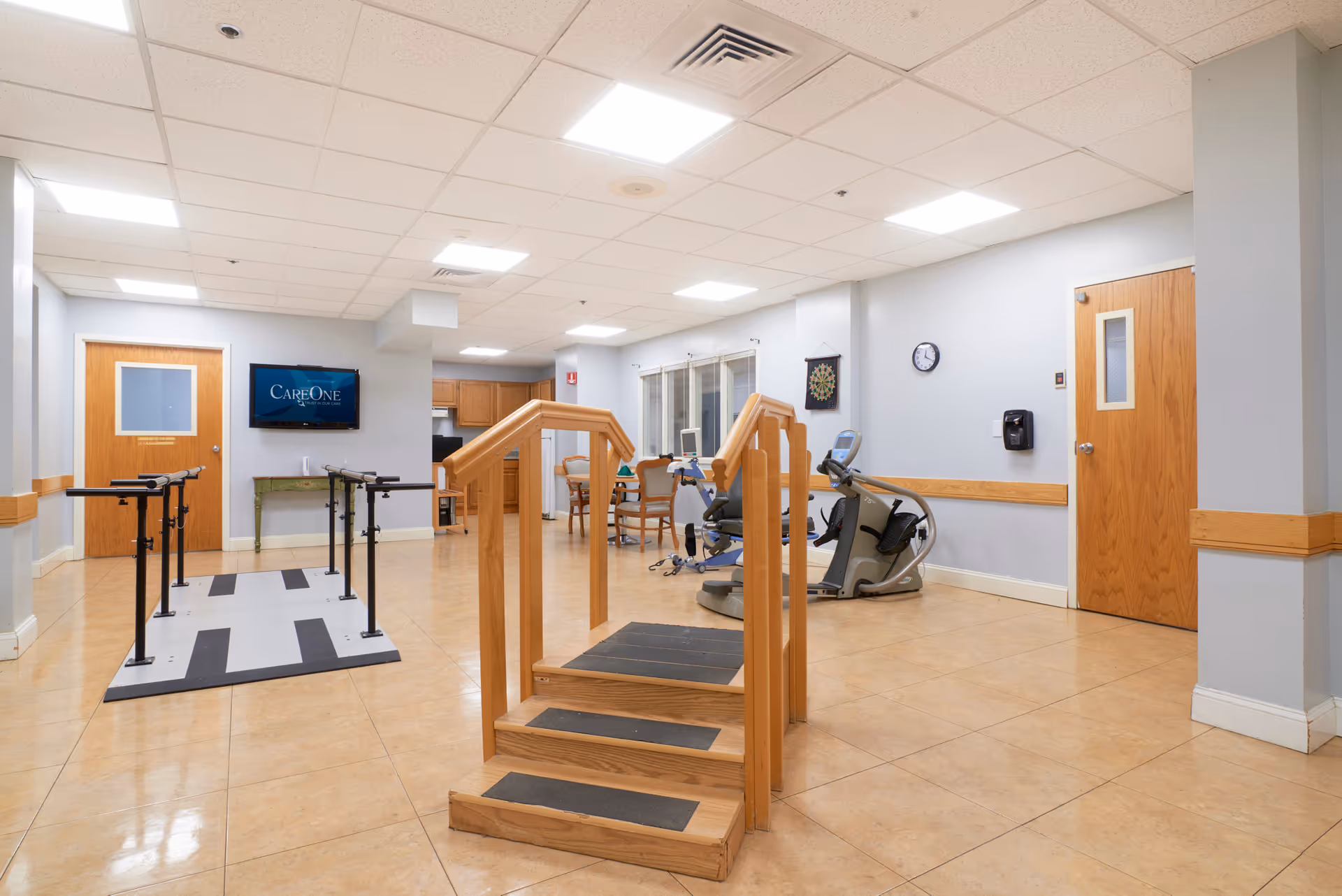 A rehabilitation or physical therapy room in a senior living facility with wooden stairs for exercise, parallel bars, exercise bikes, a dartboard on the wall, a clock, and a television mounted on the wall displaying the CareOne logo. The room has light-colored walls, tiled floors, and wooden doors.