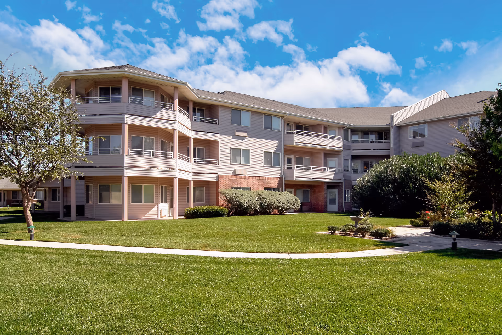 Exterior view of a three-story senior living facility building with balconies, surrounded by well-maintained green lawns, trees, and shrubs under a partly cloudy blue sky.