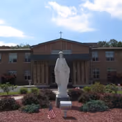 Front exterior view of a brick building with a statue of a religious figure in the foreground, surrounded by landscaping and an American flag, under a partly cloudy sky.