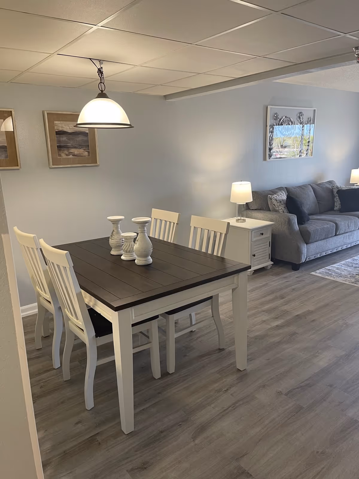 A dining area with a dark wooden table and four white chairs. Three decorative white candle holders are placed on the table. In the background, there is a gray couch with cushions, a white side table with a lamp, and framed artwork on the walls. The floor is covered with light wood laminate.