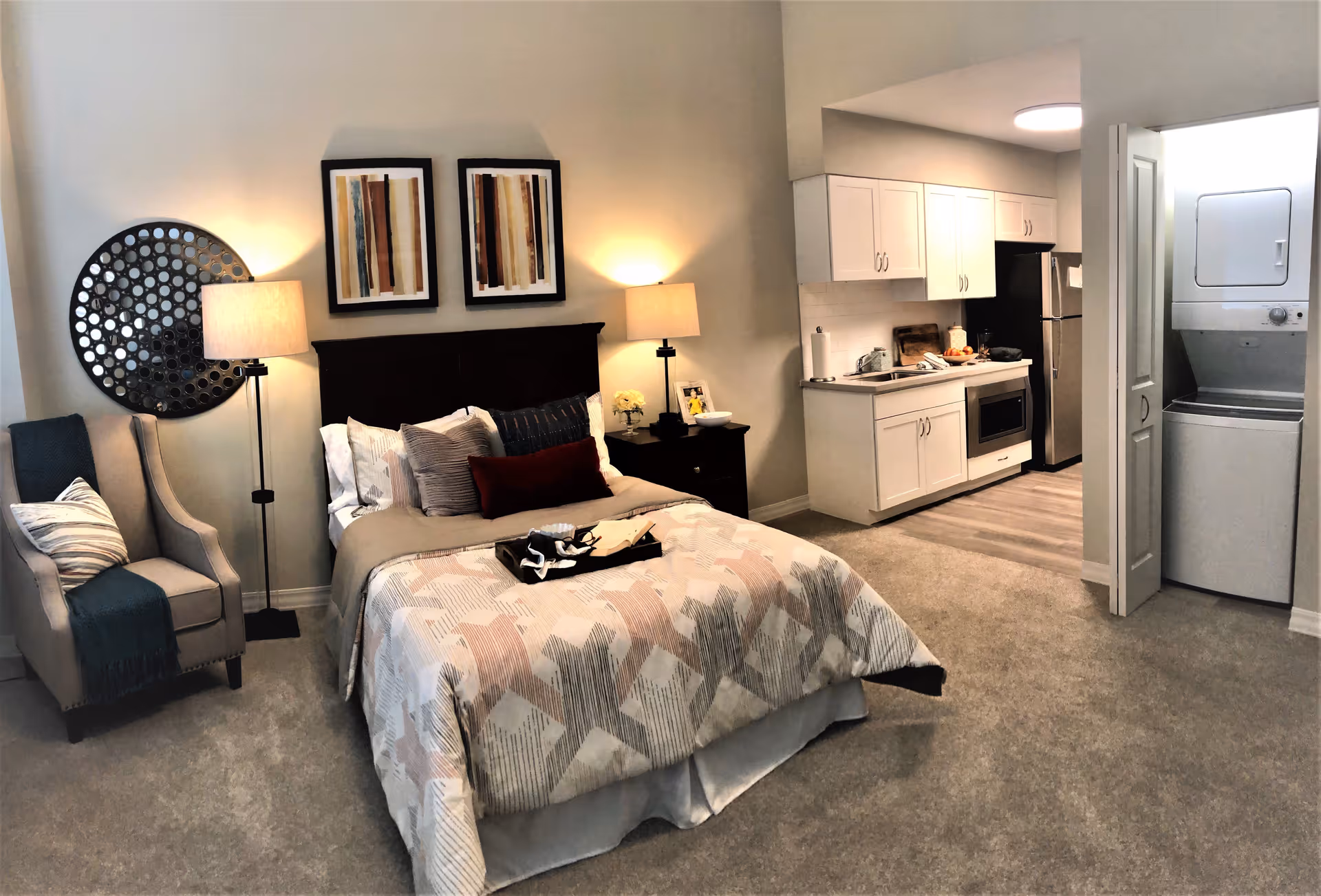 A cozy studio apartment featuring a bed with multiple pillows and a patterned comforter, flanked by two bedside tables each with a lamp. To the left of the bed is a comfortable armchair with a throw blanket and pillow. Above the bed are two framed abstract artworks and a decorative round wall piece. On the right side of the room is a small kitchen area with white cabinets, a sink, a stove, and a refrigerator. Next to the kitchen is a closet with a stacked washer and dryer. The room has carpeted flooring and soft lighting.