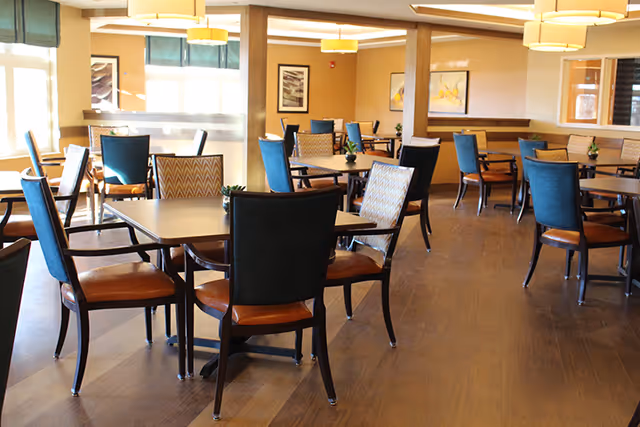 A bright and spacious dining room with multiple square tables and chairs arranged neatly. The chairs have a mix of blue and patterned upholstery with wooden legs. The room features large windows with green blinds, wooden flooring, and warm lighting from ceiling fixtures. There are framed artworks on the walls and small plants on the tables.