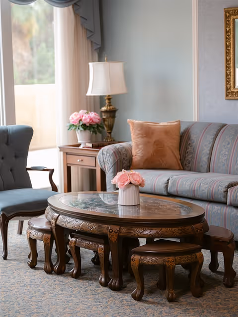 A cozy living room area with a patterned sofa featuring a peach-colored pillow, a wooden coffee table with intricate carvings and a glass top, surrounded by matching small stools. A blue upholstered armchair is positioned nearby, and a side table with a brass lamp and a vase of pink flowers sits next to the sofa. Light filters in through a window with sheer curtains.