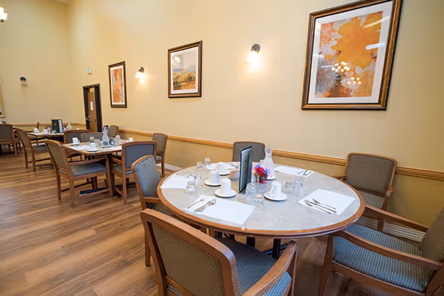 A dining room in a senior living facility with round and rectangular tables set with white cups, glasses, napkins, and silverware. The room has wooden flooring, beige walls with framed artwork, and wall-mounted lights.