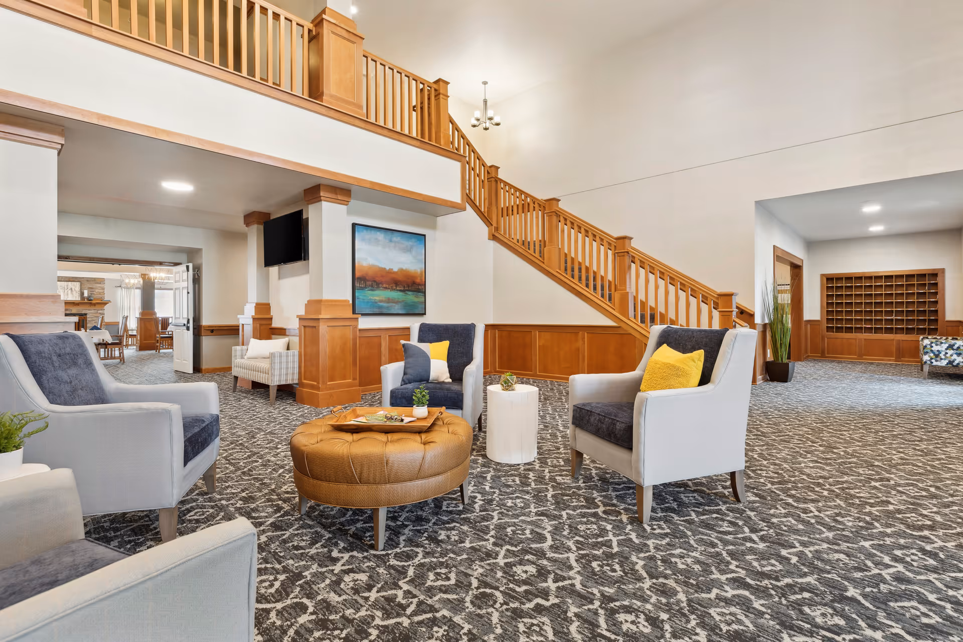 A spacious senior living facility common area with patterned carpet, four armchairs with cushions arranged around a round leather ottoman and a small white side table. There is a wooden staircase with railings leading to an upper floor, a wall-mounted TV, a large painting, and a hallway leading to other rooms with wooden paneling on the walls.