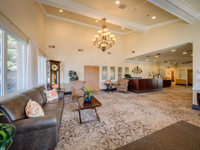 A spacious and well-lit common area with a high ceiling featuring a chandelier. The room has a patterned carpet, a brown leather sofa with floral cushions, two upholstered armchairs, a wooden coffee table with a plant, and a tall grandfather clock near large windows with white curtains. There is a reception desk in the background with decorative plants and wall art.