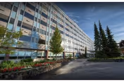 Front exterior of a multi-story senior living building with balconies, landscaped flower beds, trees, and a driveway leading to the entrance.