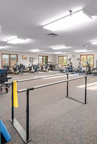A spacious fitness room with various exercise equipment including stationary bikes, elliptical machines, and parallel bars. The room has large windows letting in natural light and bright ceiling lights.