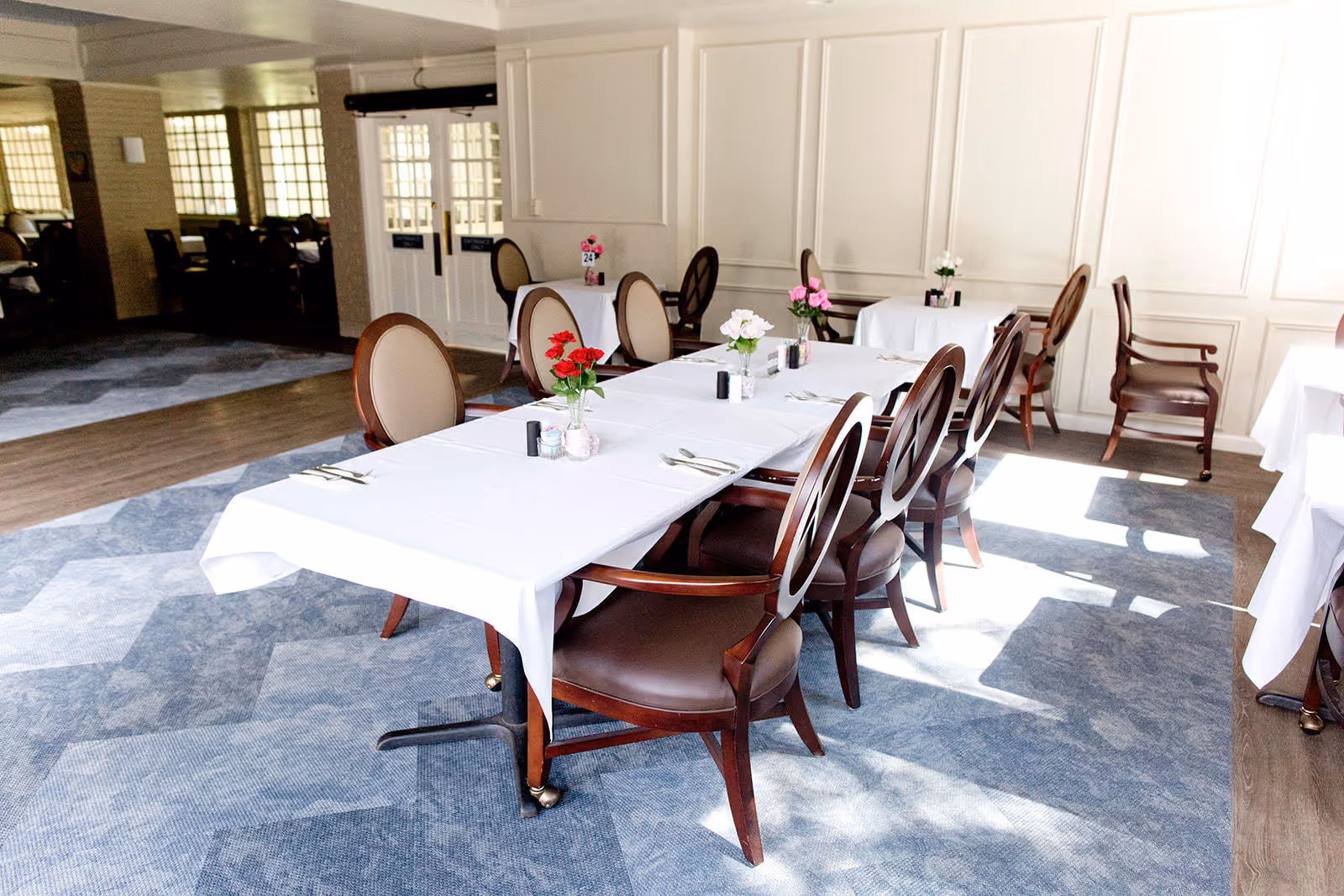 A bright dining room with tables covered in white tablecloths, each set with silverware and small vases of colorful flowers. The room features elegant wooden chairs with cushioned seats and backs, and sunlight streams in through windows, casting shadows on the patterned carpeted floor.