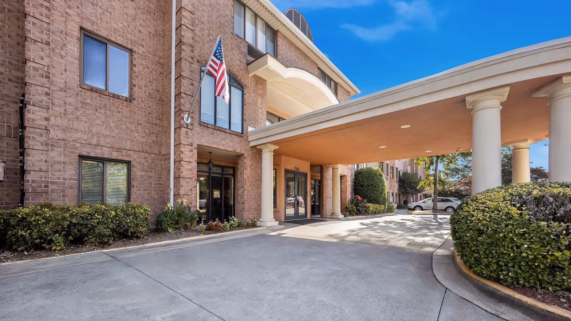 Entrance of a brick senior living facility with a covered driveway supported by large white columns. There are American and state flags mounted on the building near the entrance. Shrubs and small plants line the walkway, and a few cars are parked in the background under a clear blue sky.