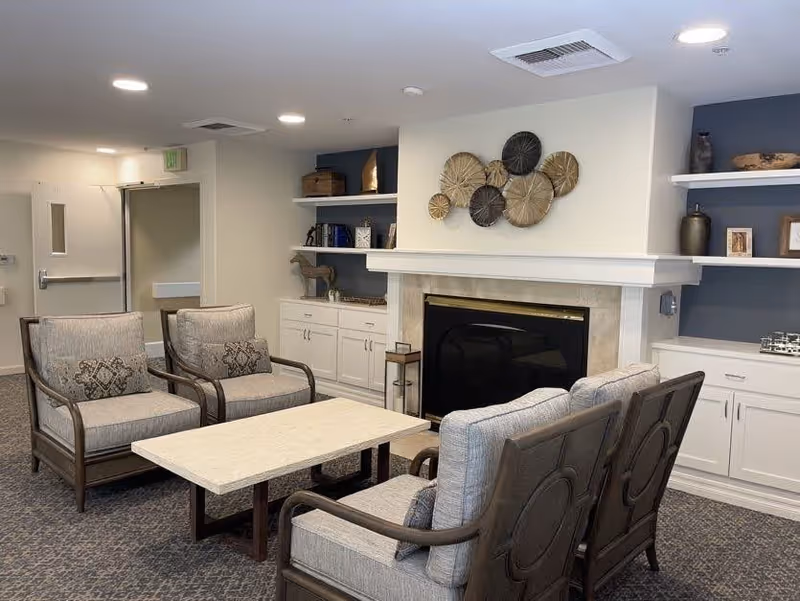A cozy living room area with four cushioned armchairs arranged around a rectangular coffee table. The room features a fireplace with decorative wall art above it, built-in white cabinets and shelves with various decorative items, and a carpeted floor. The walls are painted in neutral tones with one accent wall in blue.