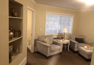 A cozy living room area with two gray upholstered armchairs and a matching ottoman. Between the chairs is a small white side table with a lamp and decorative items. A window with white blinds is behind the chairs, and built-in shelves with decorative vases and items are visible on the left side.