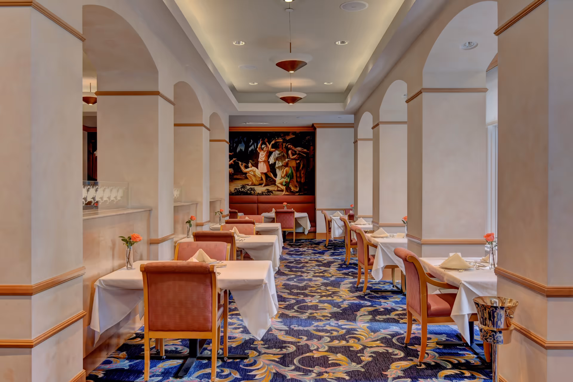 Elegant dining room with tables covered in white tablecloths, each set with napkins and a single rose in a vase. The room features arched columns, patterned carpet, and a large classical painting on the far wall. Soft lighting hangs from the ceiling.