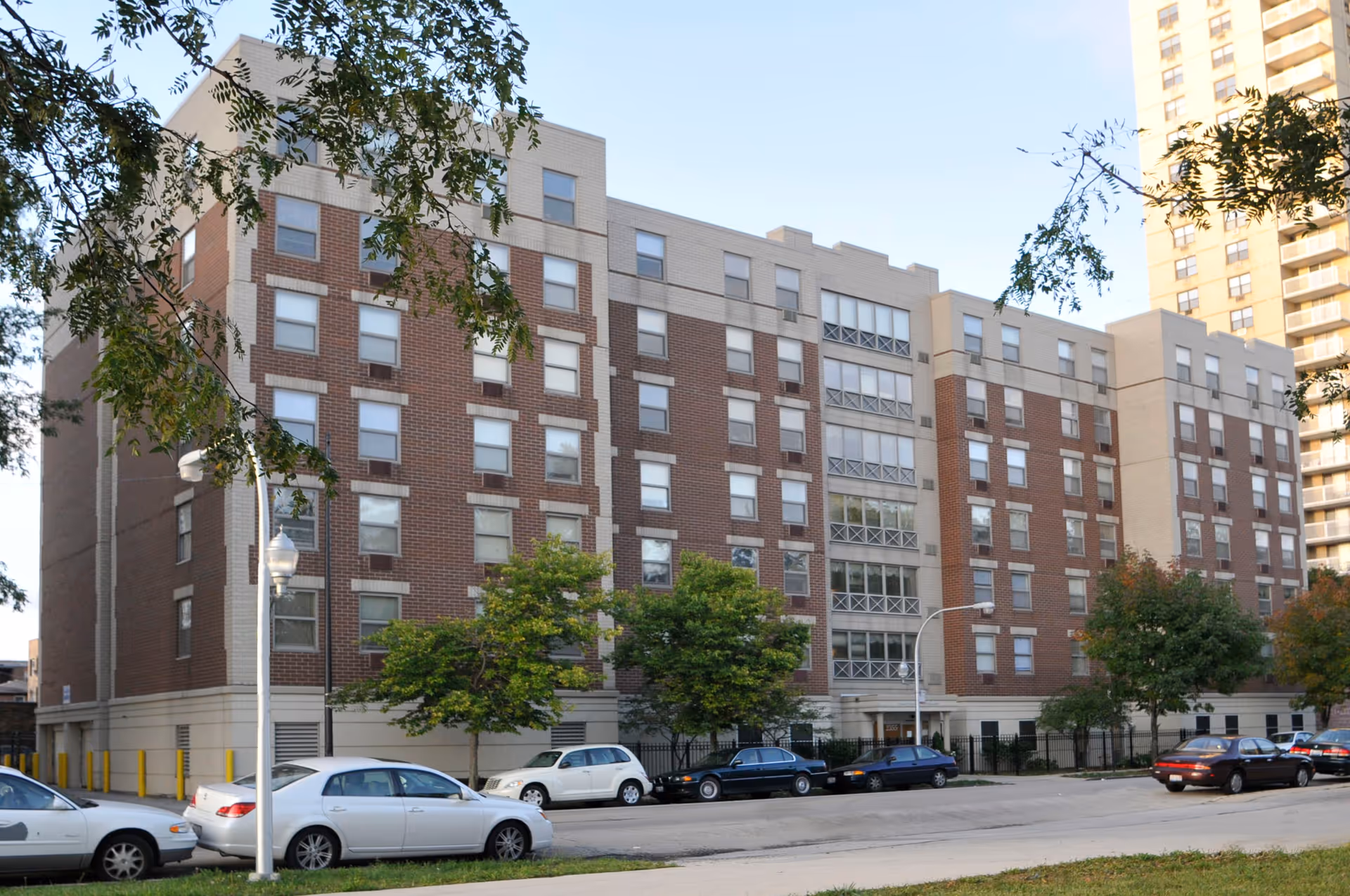 Exterior view of a multi-story brick and concrete senior living facility building with several windows, trees in front, and parked cars along the street.
