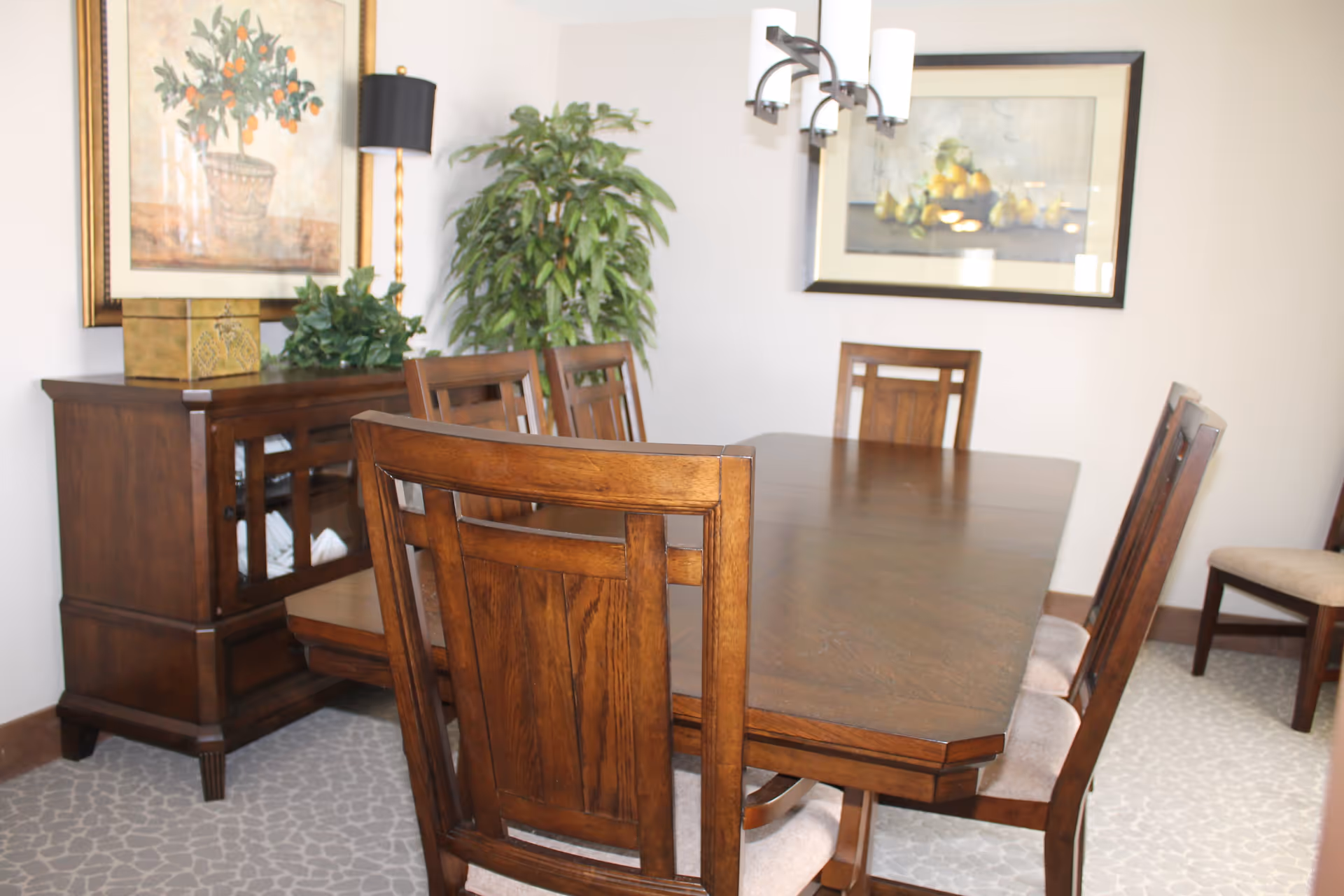 A dining room with a wooden dining table surrounded by six wooden chairs with cushioned seats. There is a wooden sideboard against the wall with decorative plants and a lamp on top. Two framed paintings hang on the walls, and a modern light fixture is mounted above the table. The floor is carpeted with a patterned design.