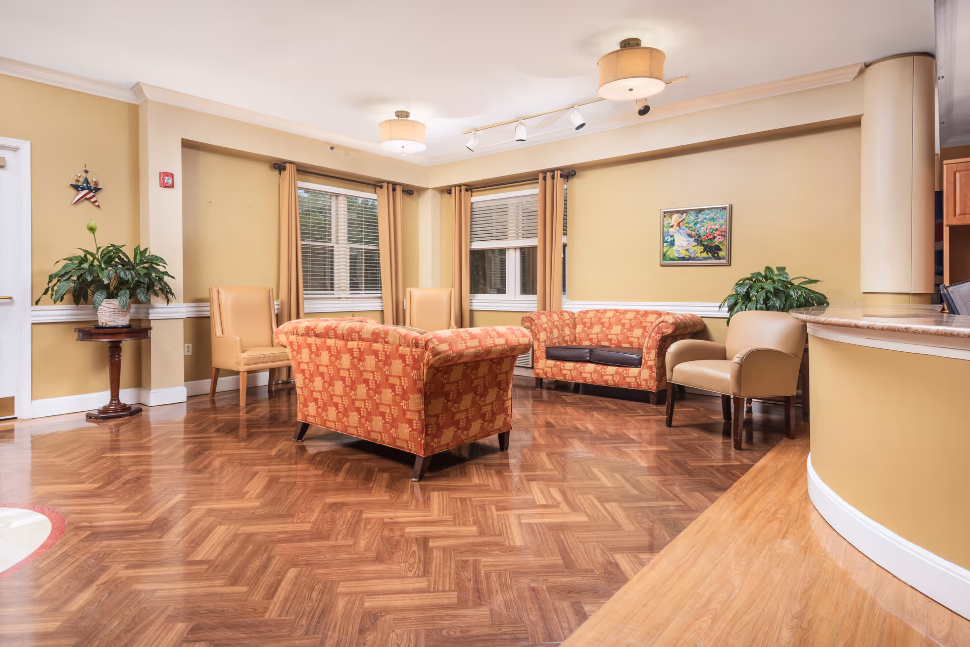 A cozy living room area with patterned orange sofas, beige armchairs, wooden parquet flooring, two windows with beige curtains, a small round wooden table with a potted plant, a colorful painting on the wall, and warm yellow walls with white trim.