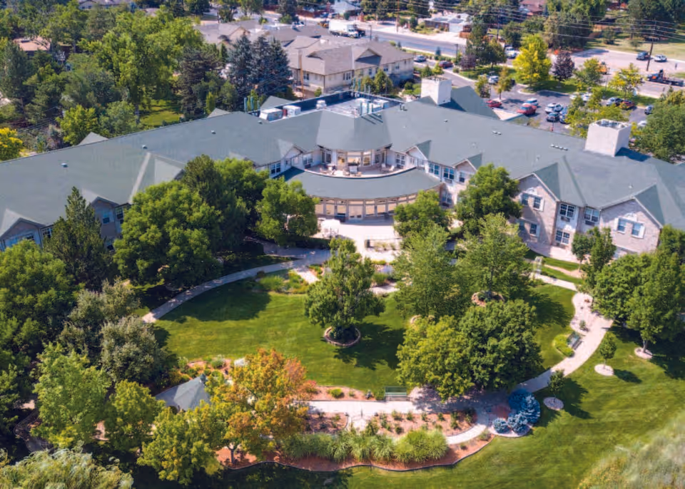 Aerial view of a senior living facility named Gardens at Columbine, showing a large building surrounded by lush green trees, landscaped gardens, walking paths, and a parking area in the background.