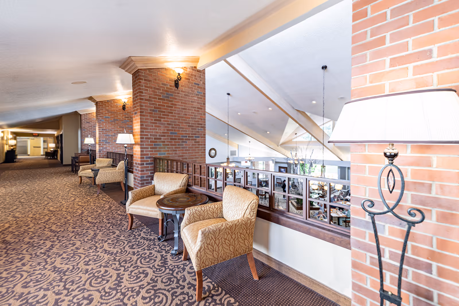 A bright and spacious hallway in a senior living facility with patterned carpet, brick pillars, and several seating areas featuring upholstered armchairs and small round tables. The hallway overlooks a large common area below with hanging pendant lights and large windows allowing natural light to fill the space.