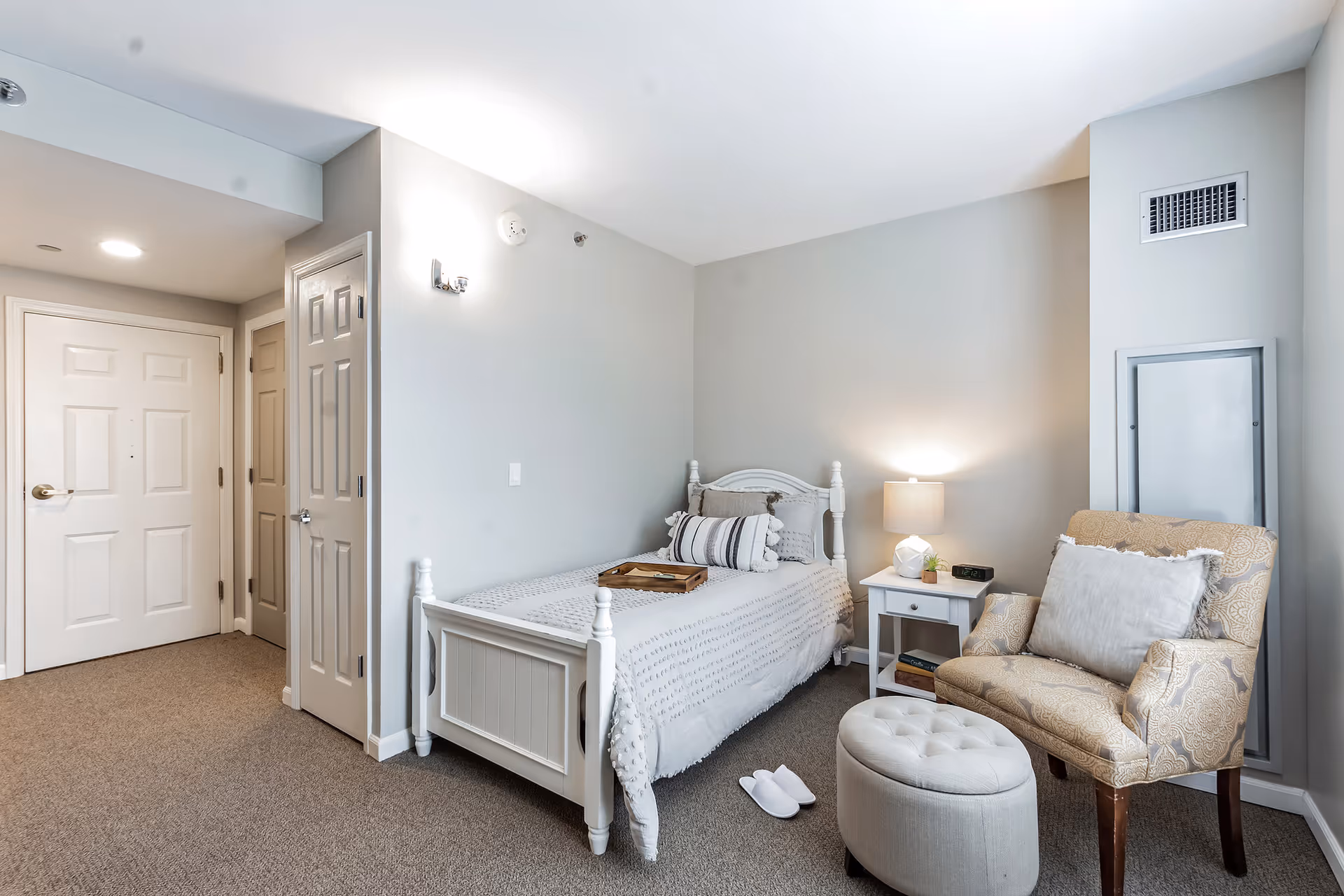 A cozy bedroom in a senior living facility featuring a single white wooden bed with a textured white bedspread and decorative pillows. Next to the bed is a white nightstand with a lamp, a small plant, and a digital clock. An upholstered armchair with a patterned fabric and a matching ottoman are positioned near the bed. The room has beige carpet flooring, light gray walls, and two closed doors near the entrance.
