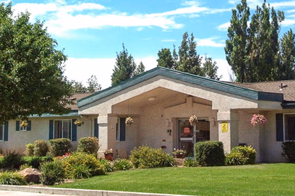 Exterior view of a single-story nursing home building with a covered entrance, surrounded by green grass, bushes, and trees under a blue sky with some clouds.