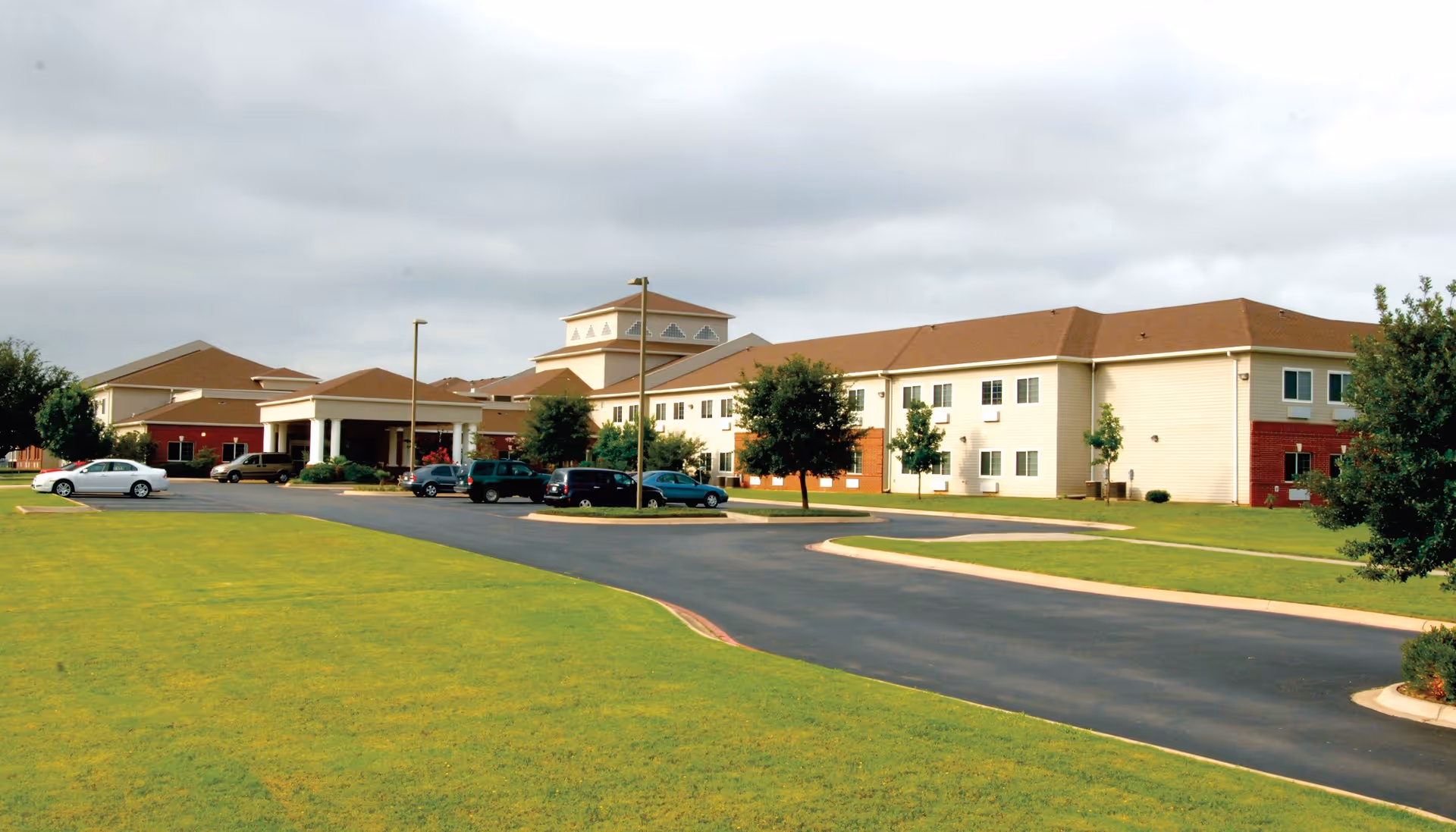 Exterior view of a senior living facility building with a driveway, several parked cars, green lawns, and trees under a cloudy sky.