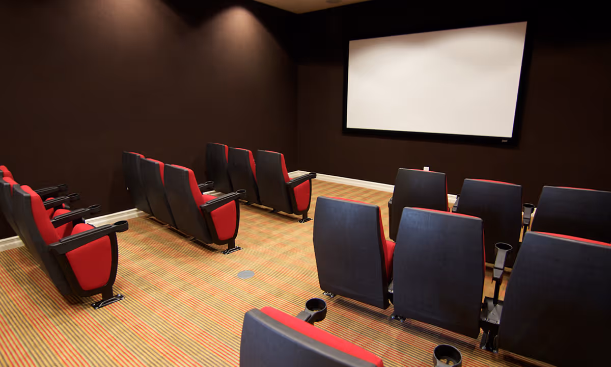 Small theater room with rows of black and red cushioned seats facing a large blank white projection screen on a dark brown wall. The floor is carpeted with a striped pattern in shades of orange and beige.