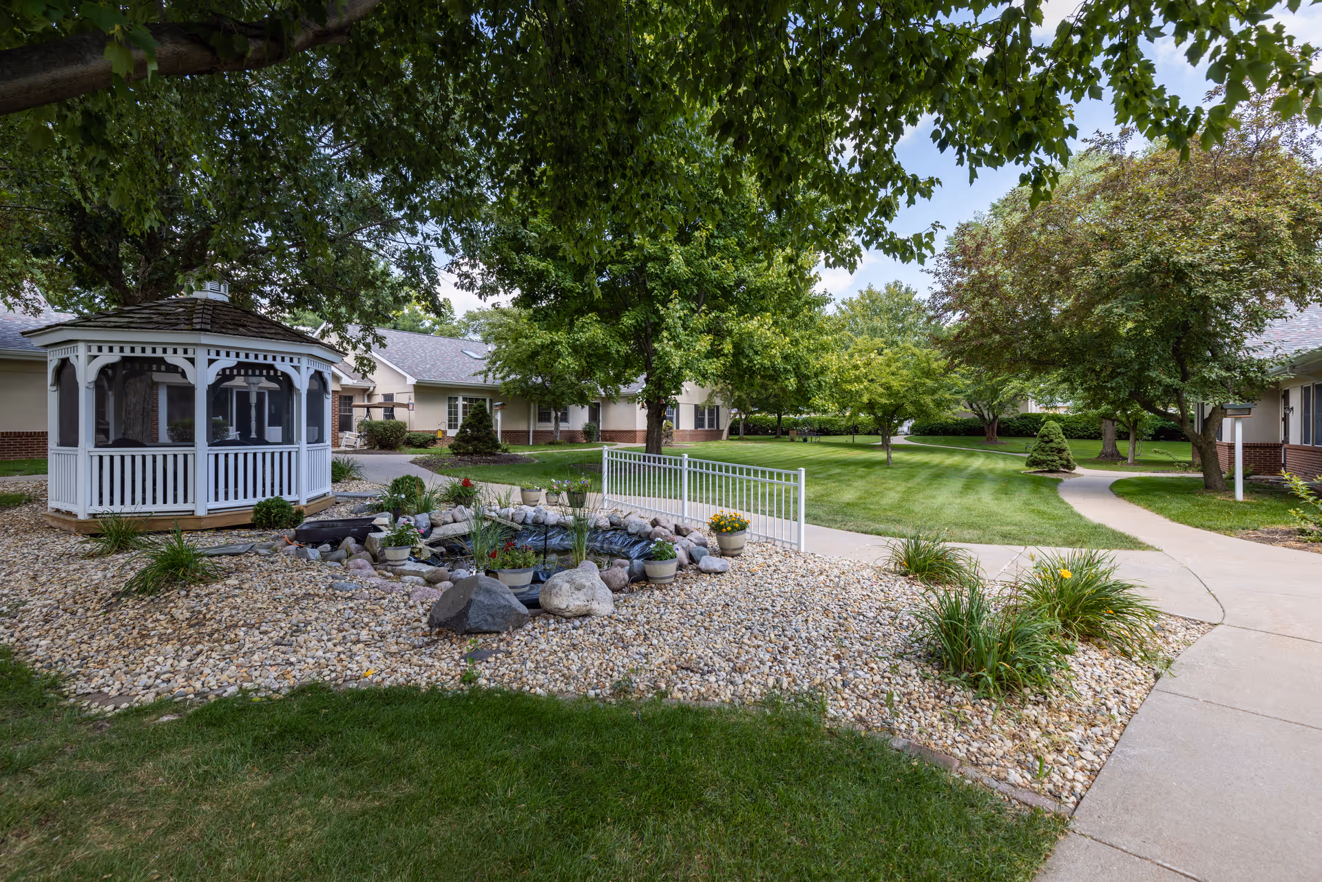 A peaceful outdoor garden area at Country Manor Memory Care featuring a white gazebo, a small pond surrounded by rocks and plants, well-maintained green lawns, trees, and paved walkways leading to single-story buildings in the background.