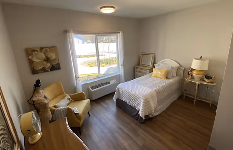 A bright and cozy bedroom with a single bed covered in a white and gray striped quilt, a yellow pillow with the word 'happy', a small bedside table with a lamp and decorative items, a yellow armchair with a pillow and a throw blanket, a window with white curtains letting in natural light, a dresser with a fan and a mannequin, and a painting of a flower on the wall.