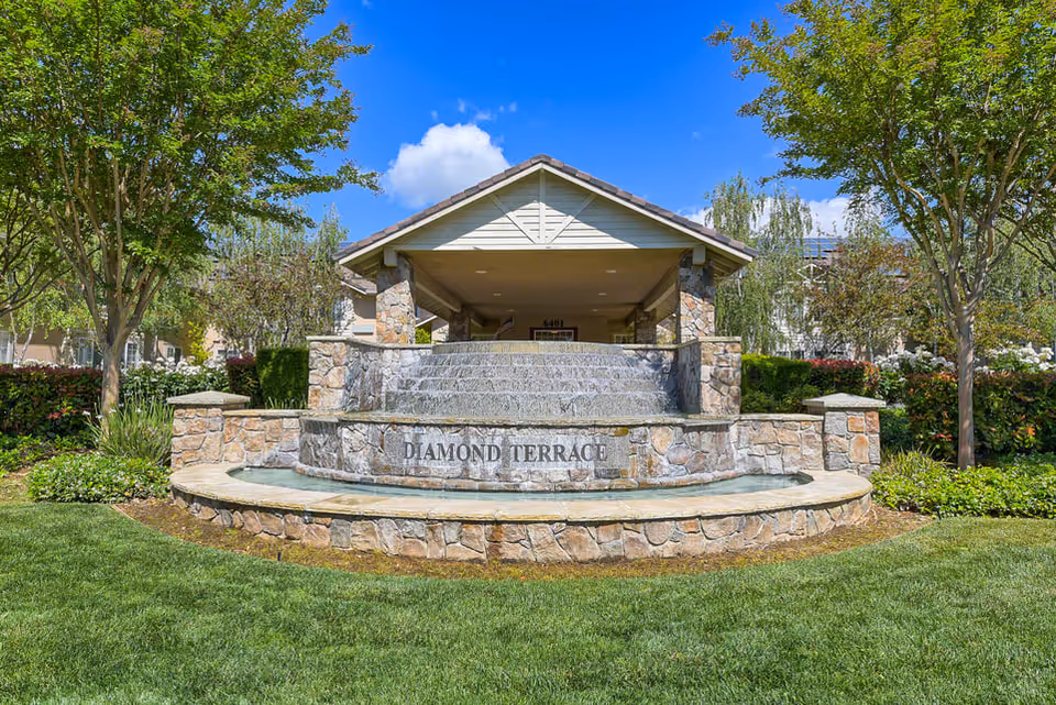 Stone water fountain with cascading water in front of a covered entrance structure, surrounded by green grass, trees, and shrubs under a blue sky with some clouds. The fountain has the words 'DIAMOND TERRACE' engraved on it.