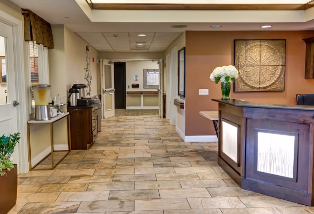 Interior view of a senior living facility hallway with a reception desk on the right, decorated with a vase of white flowers and a framed decorative panel on the wall. On the left side, there is a table with a beverage dispenser and coffee station. The floor is tiled with a beige and brown pattern, and the hallway leads to open doors at the far end.