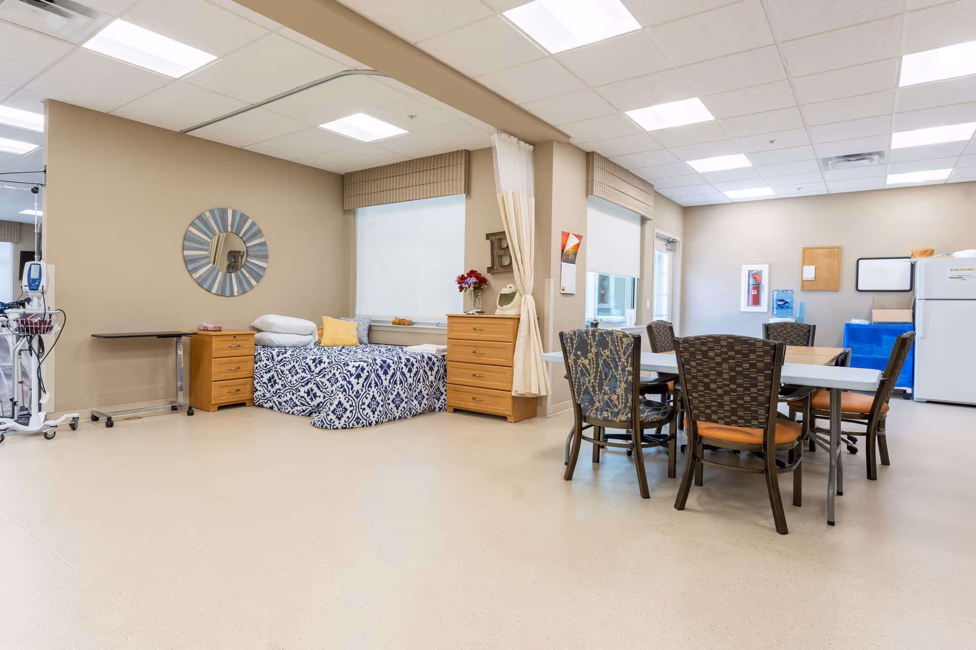 Bright multipurpose room in a senior care facility with a single bed, dressers, and a dining table with chairs.