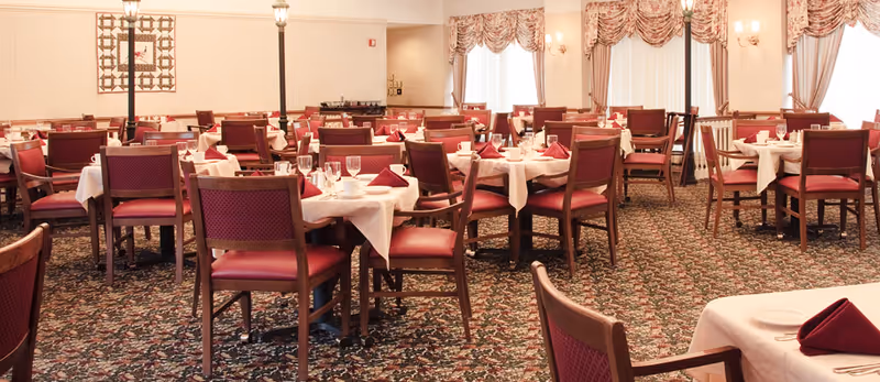 A spacious dining room with multiple tables covered in white tablecloths, each set with folded red napkins, glassware, and plates. The room features patterned carpet flooring, wooden chairs with red cushions, large windows with draped curtains, and soft lighting from wall sconces and standing lamps.