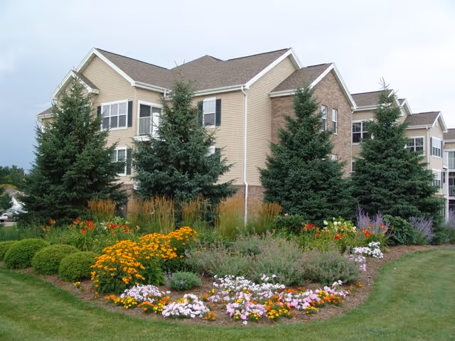 Beige multi-story senior living building with evergreen trees and colorful flower beds in the foreground.